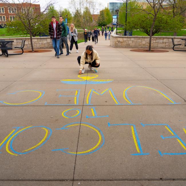A student writes "Welcome to UWEC" in chalk with students walking behind her.