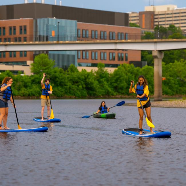 Students paddle board on the Chippewa River with the newly-constructed Science and Health Sciences building in the background.