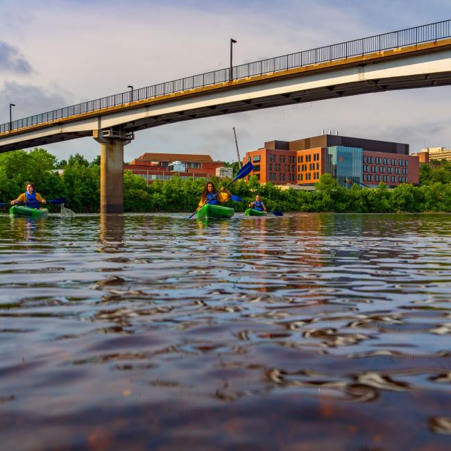 Students kayak on the Chippewa River with the Science and Health Sciences building and the bridge in the background.