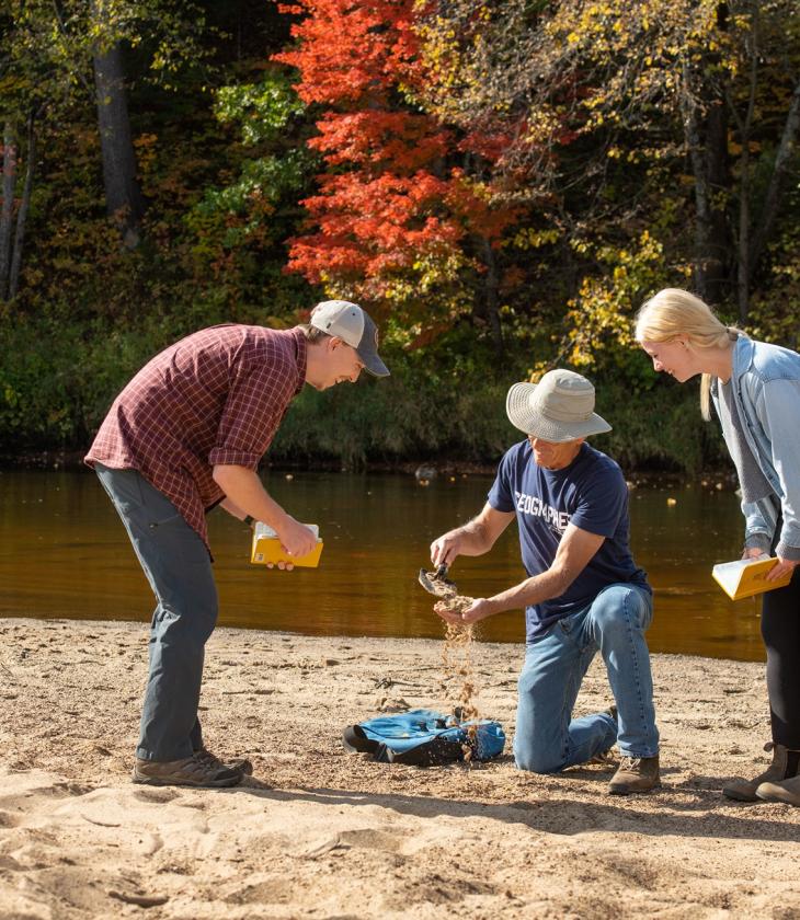 A professor kneels down and pours sand into his hand while two students smile and observe
