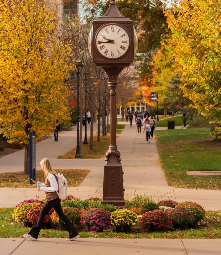 Student walks across campus in autumn in front of clock.