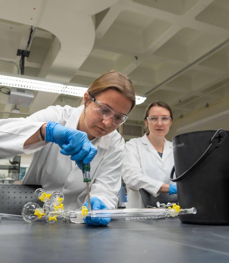 Two students wearing white lab coats work together connecting a piece of tubing in a lab setting 
