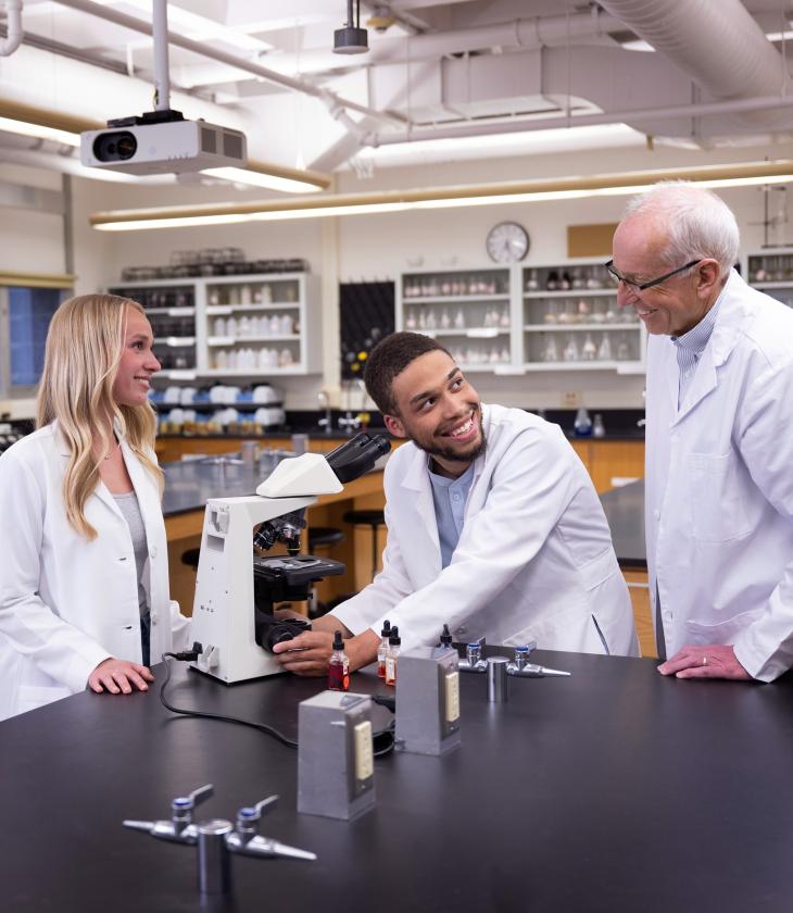 Students and a professor in white lab coats work together in a lab, smiling.