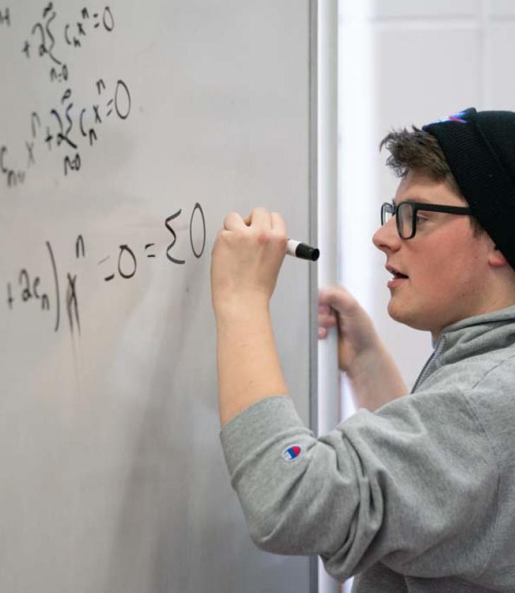 Student in Blugold sweatshirt solves a math problem on a whiteboard.