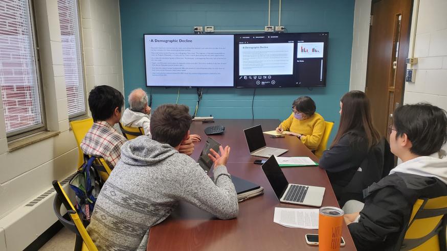 Students sit around a table and look at two monitors mounted on a wall in a student research room on campus
