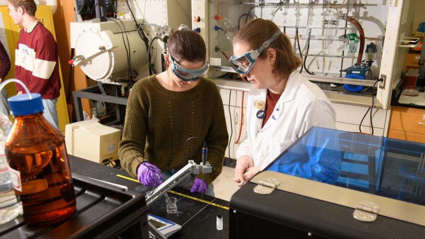 two women in chemistry lab, goggles, one in white lab coat
