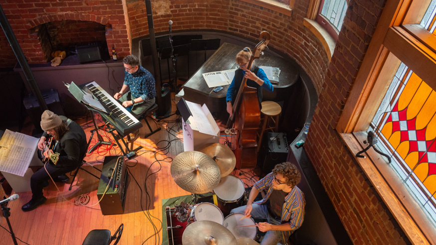 A jazz band performing in a cozy, brick-walled venue with stained glass windows, featuring musicians on keyboard, guitar, double bass, and drums.