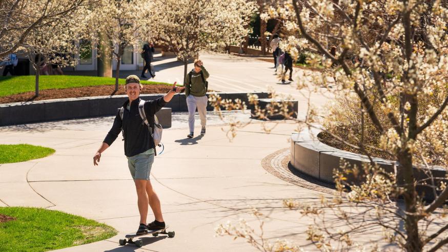 male student on a long board crossing campus