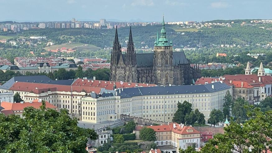 An aerial view of Prague showing the historic Prague Castle with its Gothic spires, surrounded by modern cityscape and lush greenery.