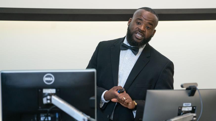 Man wearing a black suit and bowtie speaks in front of two computer monitors