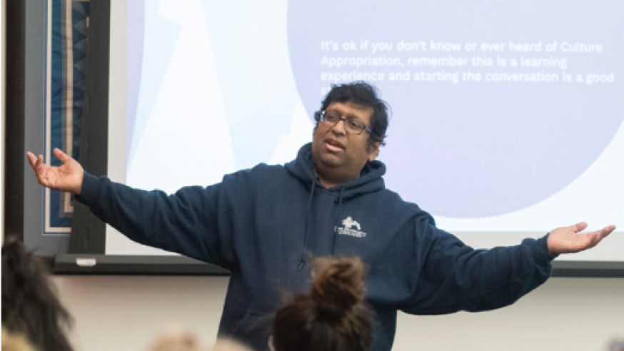 Professor stands in front of projector screen with arms outstretched while talking.
