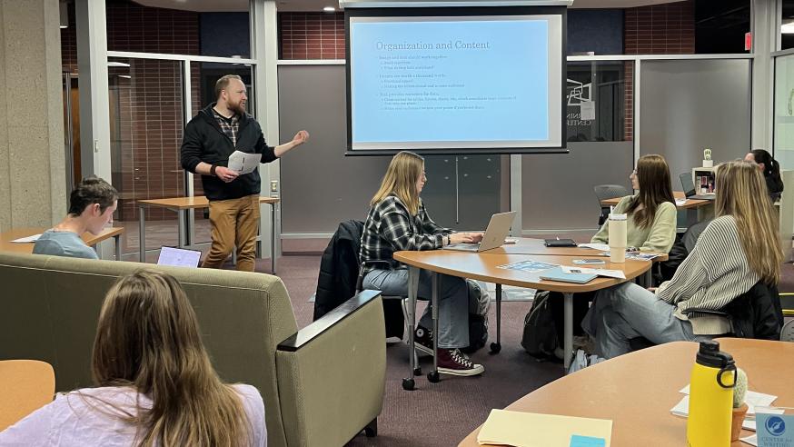 A glass room in the library. Students are sitting on couches and at tables while a male-presenting person speaks in front of a projector screen.