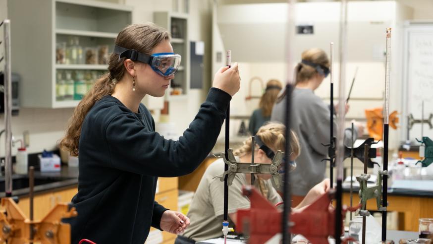 A student wearing safety goggles examines lab equipment during a Chemistry lab