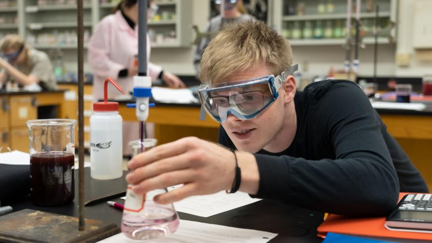 Student wearing safety goggles examines a beaker with liquid during a Chemistry Lab