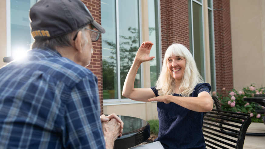 Woman smiles while signing using ASL in conversation with another person.