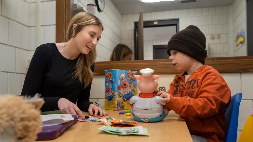 A student clinician works with a child at a table, talking about the child's toy.