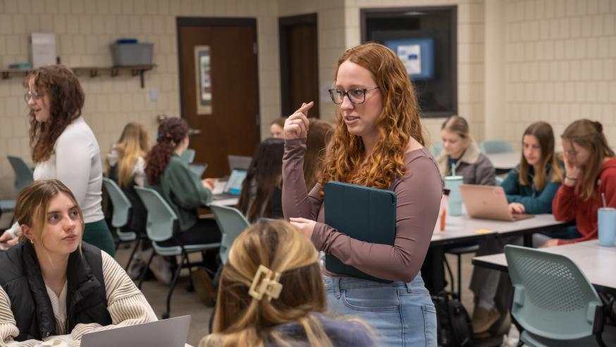 A student stands up while speaking to a group of students seated at a table.
