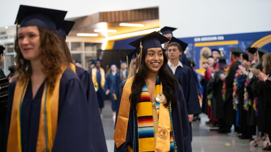 Students wearing graduation caps and gowns smile as they walk in to the ceremony