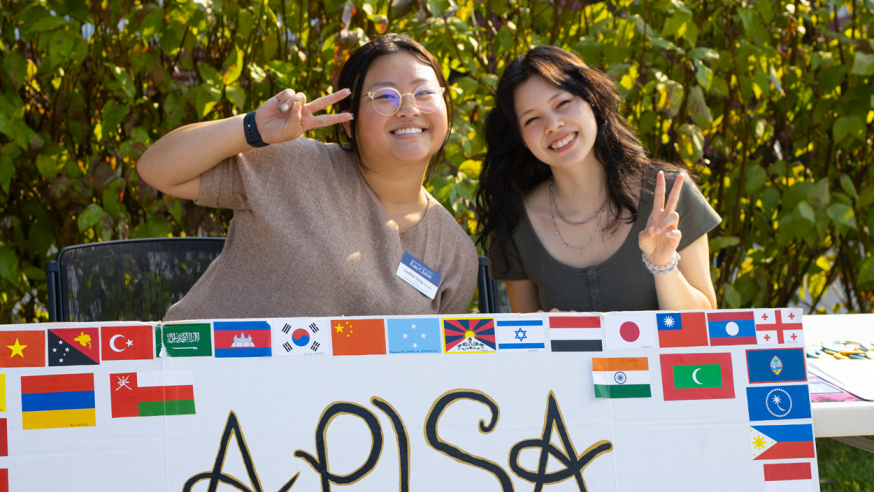 Two students smile at the camera with an 'APISA' sign in front of them