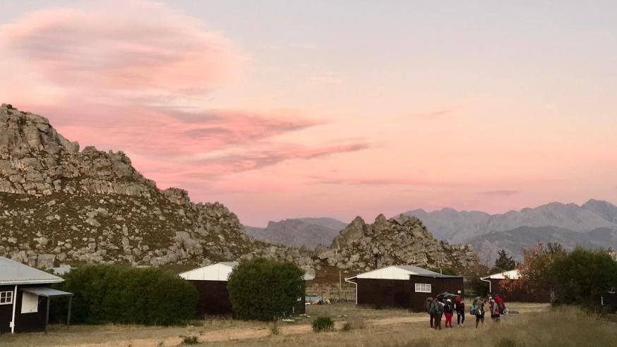 People hiking in front of mountains under a pink sunset sky
