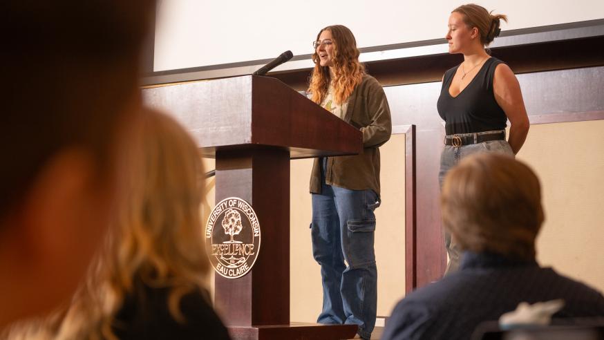 Two students speak at a podium to a crowd.
