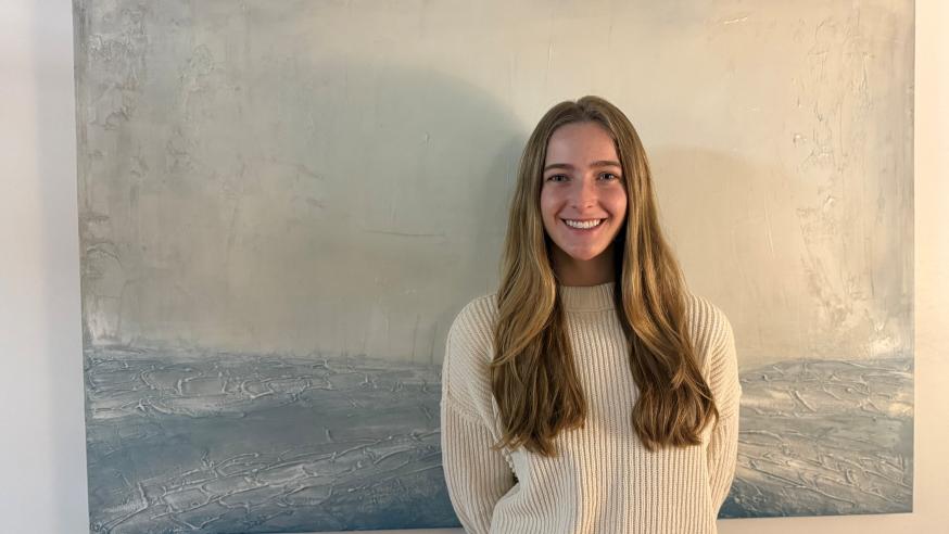 UWEC student Maren Durand stands smiling in front of a subtle blue and white waterscape painting.