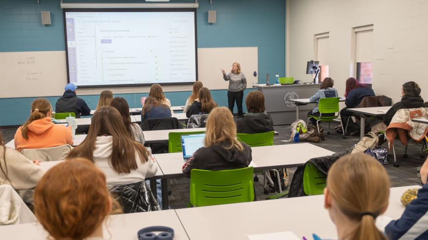 A professor speaks to a class while gesturing at a PowerPoint screen