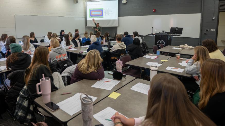 A professor stands in front of a PowerPoint while speaking to a full classroom of students