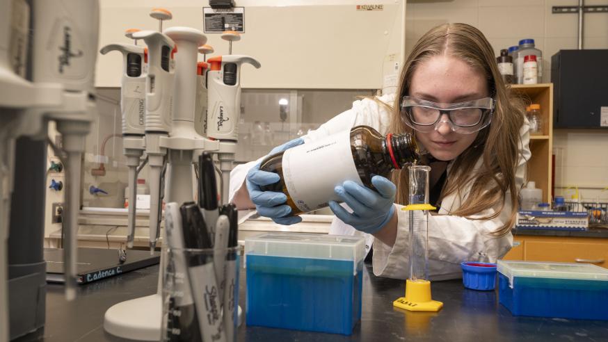 A student pours liquid into a glass beaker while wearing a lab coat, gloves, and safety goggles.