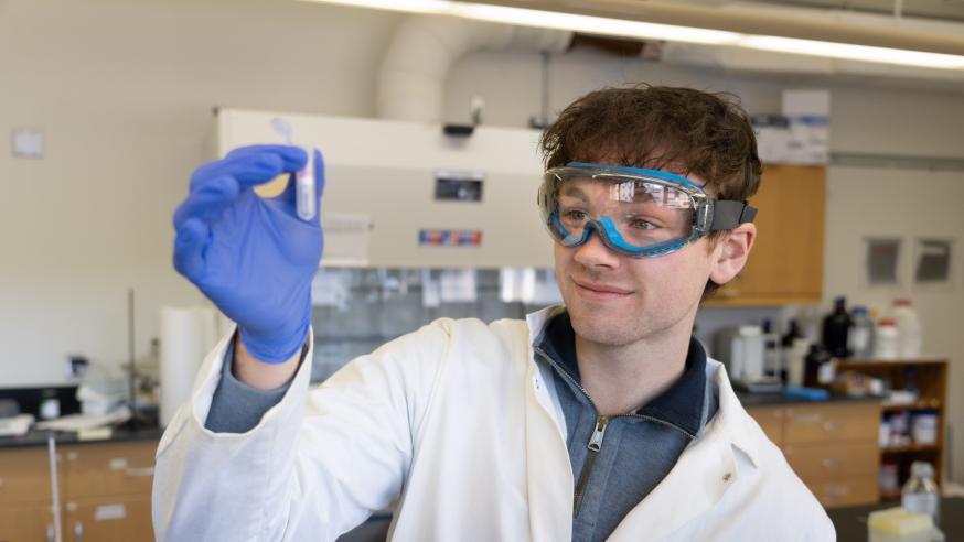 An Integrated Studies student examines a small vile while wearing gloves, safety goggles, and a white coat.