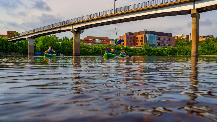 Students kayak on the Chippewa River with the Science and Health Sciences building and the bridge in the background.