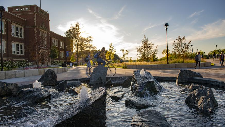 Students walk and bike next to the Stowe Fountain during a sunset on campus