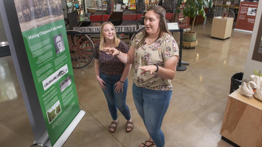 UW-Eau Claire public history graduate students Jordan Stish (left) and Alexi Linder discuss the exhibit they helped create in the Irvine Park Welcome Center in Chippewa Falls. (Photo by Shane Opatz)