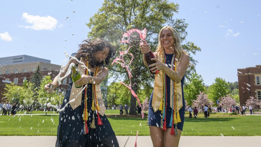 two women spraying bubbly outside on a sunny day, graduation gowns.