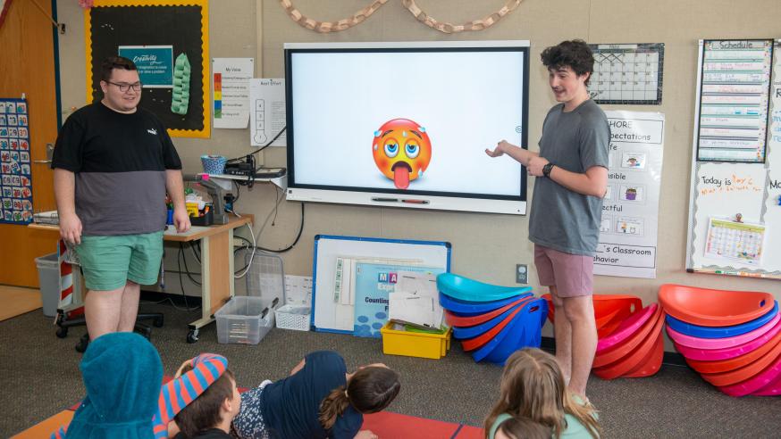 male teaching students at a board in front of elementary students, young kids all seated on the floor
