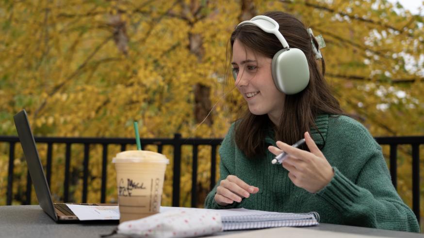 female student on the campus patio at a tble using laptop and headphones, smiling