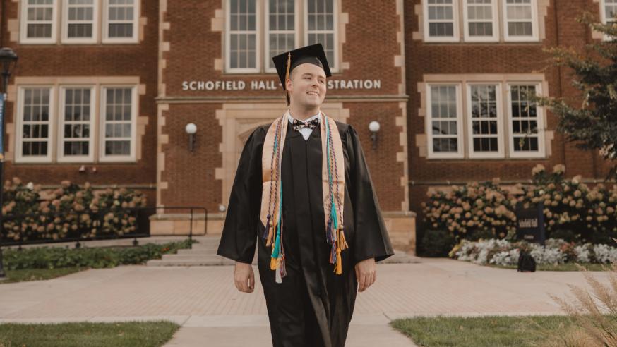Jordan Blue smiling  in cap and gown in front of Schofield Hall, summer or fall sunset hour