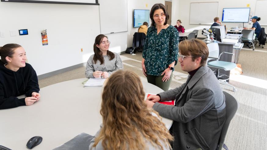 Students in a bright classroom seated at tables, instructor standing next to one group of 4.