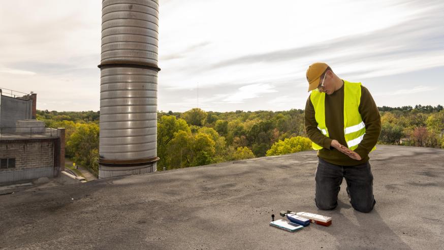 A male student wearing safety gear kneeling on a metal rooftop checking some measurement instruments