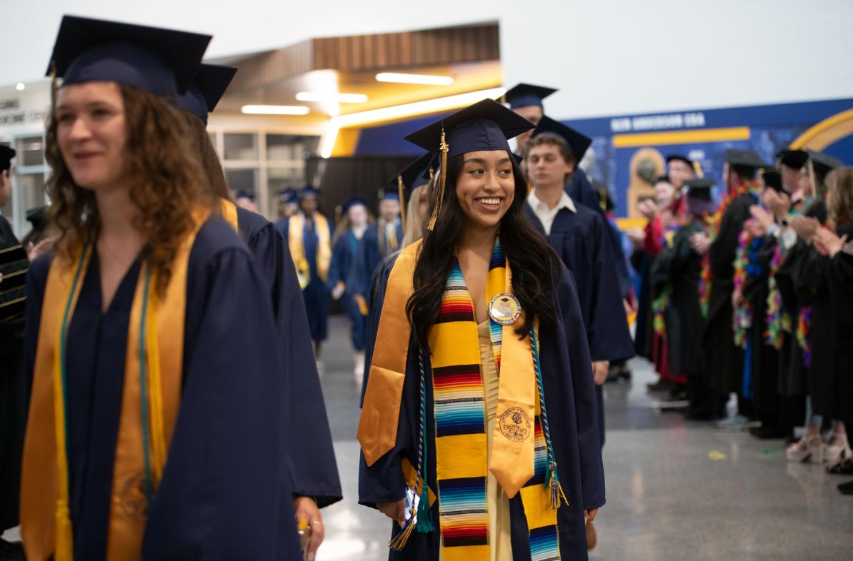 Students wearing graduation caps and gowns smile as they walk in to the ceremony