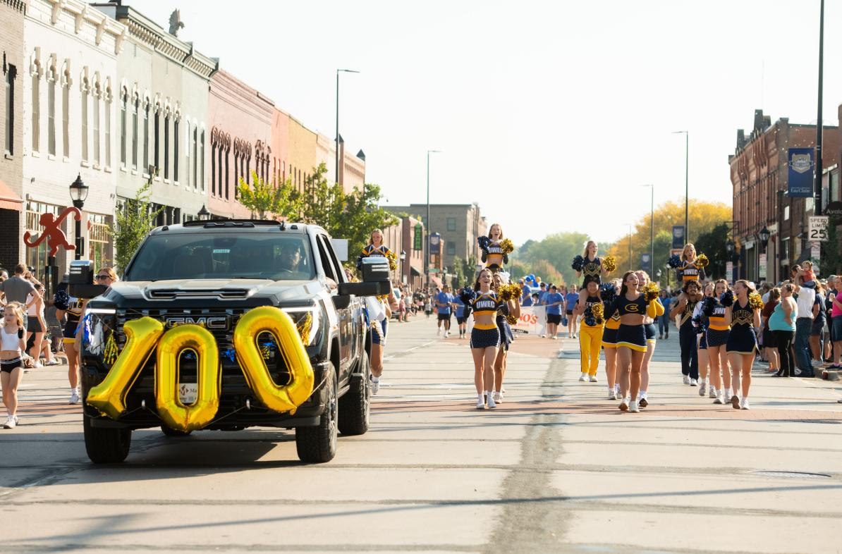 A fun view of the Homecoming parade featuring a car with the number "100" in balloons on the front while people march alongside.