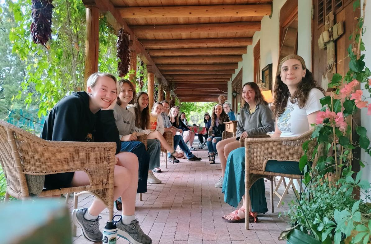 New Mexico immersion students sit across from each other, down a long, covered porch, with plants and greenery around