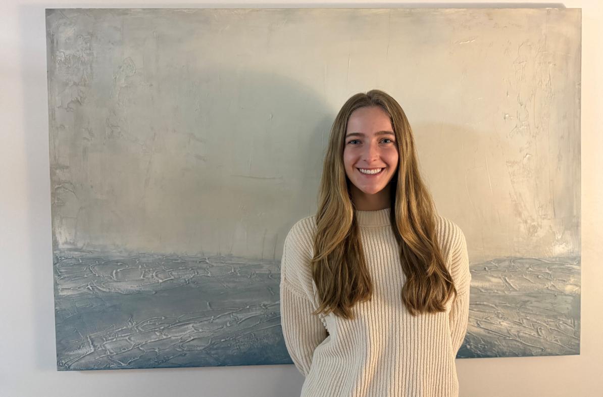 UWEC student Maren Durand stands smiling in front of a subtle blue and white waterscape painting.