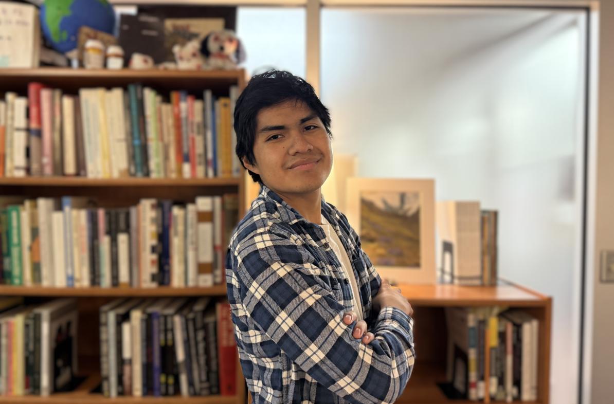 Honors student Kevin Miro stands smiling with arms crossed in front of bookshelves