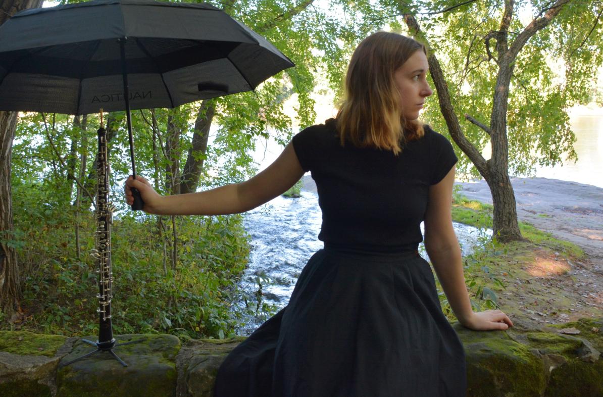 Carrie Schwartz in a black dress in the forest holds an umbrella over an oboe 
