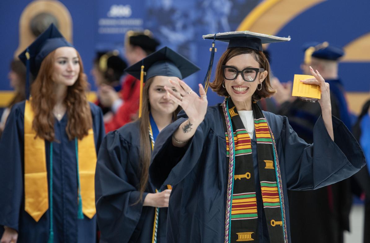 Students celebrate at graduation. One student smiles brightly and waves to the crowd.