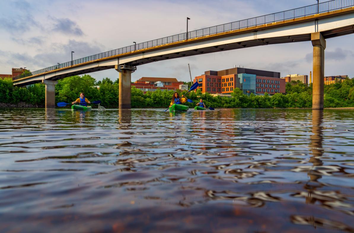 Students kayak on the Chippewa River with the Science and Health Sciences building and the bridge in the background.