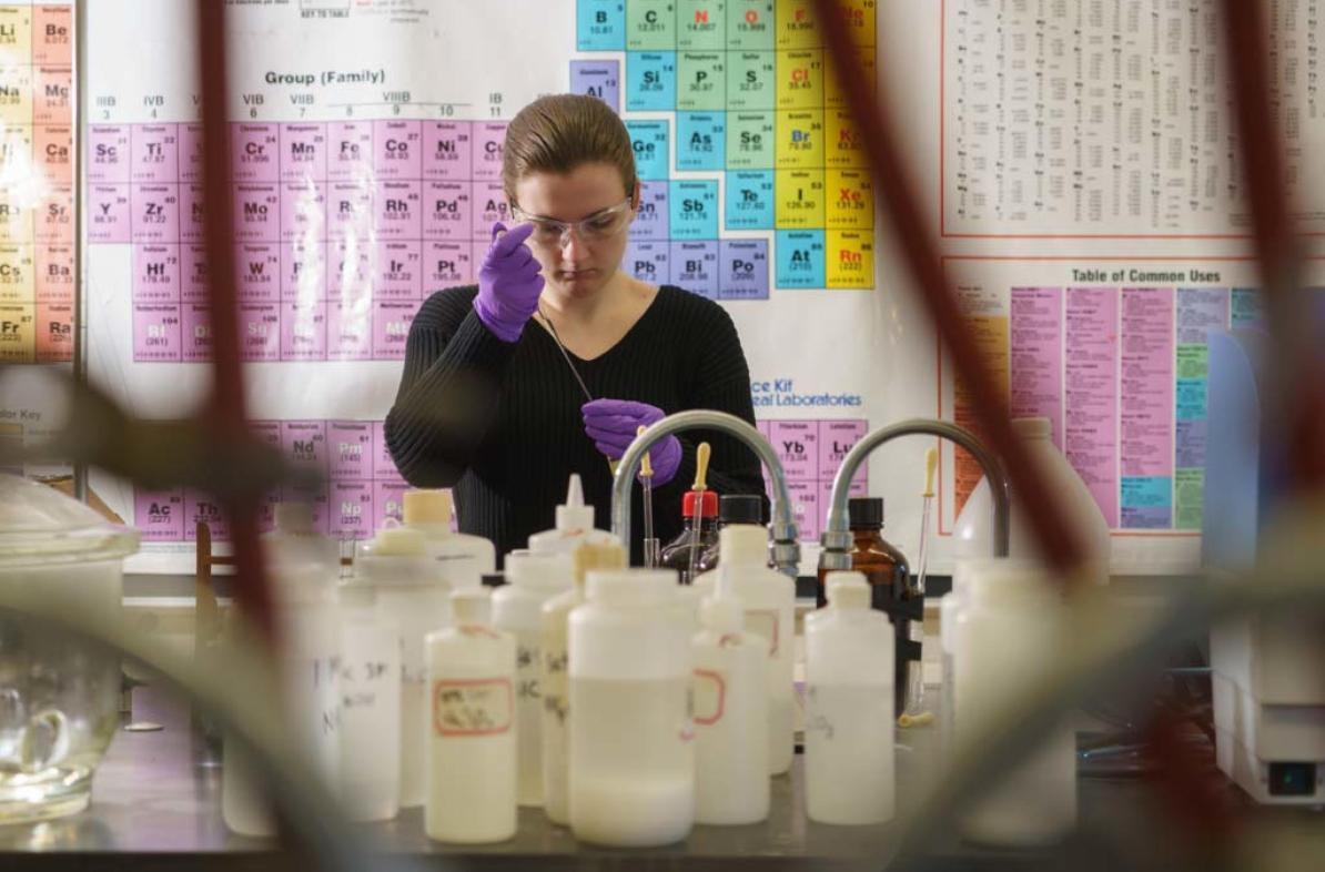 A student works with lab instruments with the periodic table hanging on the wall behind.