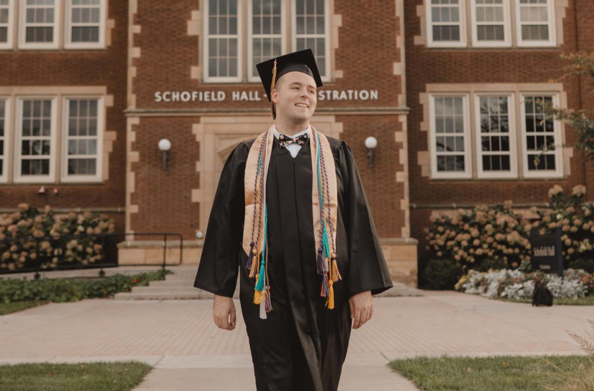 Jordan Blue smiling in cap and gown in front of Schofield Hall, summer or fall sunset hour