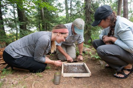 Group of researchers examining a soil sample in a forested area, using a wooden frame and metal can, while kneeling on the ground and focusing on their outdoor fieldwork.